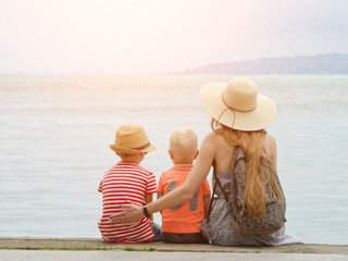 Mom and two sons sit on the pier and admire the sea and the mountains in the distance. Back view
