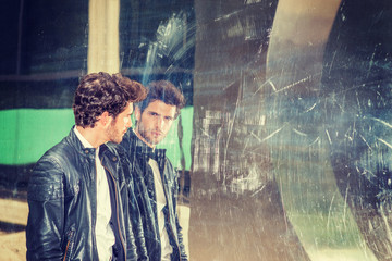 Man looking at mirror. A young handsome guy with beard, walking by metal mirror wall, looking at the reflection. Concept of self assured, self esteem, self checking strategies. .