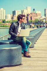 European businessman traveling, working in New York. Wearing black leather jacket, blue jeans, brown boot shoes, sunglasses, a guy with beard, sitting on bench at harbor, working on laptop computer..