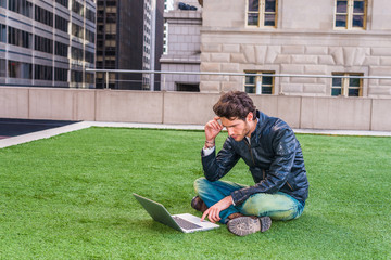 European graduate student studying in New York. Wearing black leather jacket, blue jeans, boot shoes, young guy with beard, crossing legs, sitting on green lawn, reading, working on laptop computer.