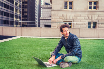 Obraz premium European graduate student studying in New York. Wearing black leather jacket, blue jeans, boot shoes, young guy with beard, crossing legs, sitting on green lawn, working on laptop computer, thinking..