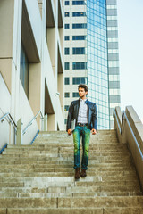 Young professional with beard, wearing leather jacket, jeans, arm carrying laptop computer, walking down stairs outside office high building after busy working day, going home. .