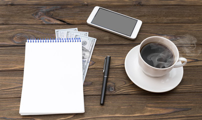 Cup of coffee, smartphone, dollars, notepad and pen on wooden desk.