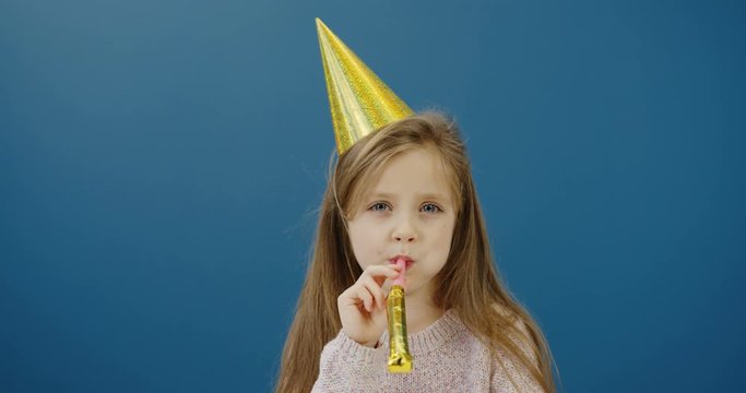 Close up of the cute pretty little girl in a birthday hat blowing in the party horn and looking straight into the camera on the blue wall background. Portrait. Inside