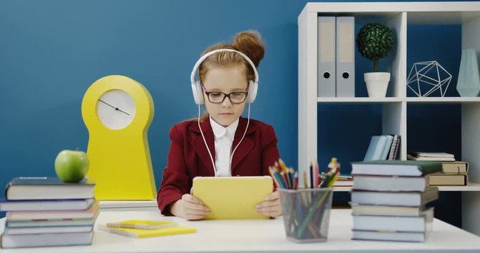 Nice schoolgirl in the glasses sitting at the desk with textbooks and listening to the music on her tablet throught headphones. School class. Indoor
