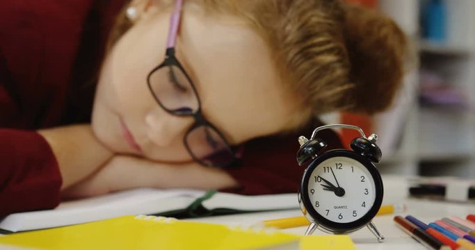 Lovely tired schoolgirl sleeping on the textbook at the table with school stuff. Alarm clock in the front. Close up. Indoor