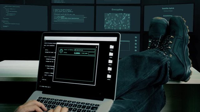 Hacker feet up to table. Hacker with laptop coding exploit to crack code. Man in military shoes sits and feet up on desk. Laptop computer screen. Monitor room dark night many screens lights.