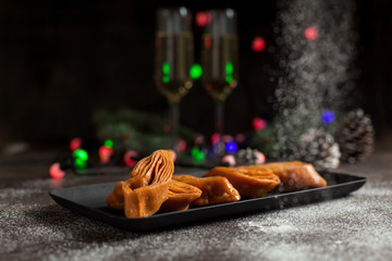 New Year's still-life. Glasses with champagne, New Year's lights, Christmas tree and sweet pastry. On a black plate