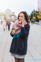 Fototapeta premium young beautiful brunette girl with coffee to go and smartphone on the background of blurred city street