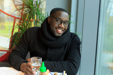 Young afro american man in glasses drinks water, sitting in a cafe.