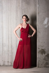 Vertical portrait of a beautiful young woman in long red dress in full growth on a background of gray concrete walls