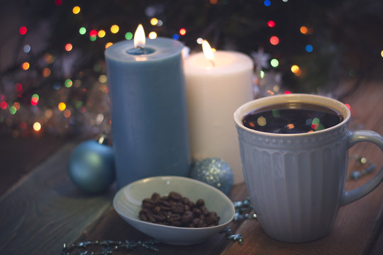 A romantic festive still life with burning candles and a bowl of coffee beans. A cup of freshly brewed coffee in a close up. Ligth blue colors. Dark Christmas background. Blurred bokeh