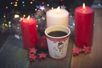 Obraz na płótnie Canvas A romantic festive still life with burning candles and a paper cup of coffe with Christmas design. A colored wooden surface. White and red colors. Dark Christmas background. Blurred bokeh