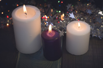 A purple and two white candles of different size in a row. A colored wooden surface. Dark Christmas background. Blurred bokeh