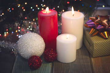 A festive still life with a red and two white candles of different size, a handcrafted gift box with a purple and orange shiny bow, white and red shiny balls. Dark Christmas background. Blurred bokeh