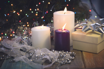 Fototapeta na wymiar A festive still life with a purple and two white candles of different size, a beige gift box with a silver bow and a white and silver garland of a ribbon and beads. Dark Christmas background. Blurred