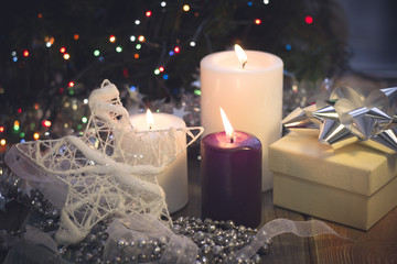 A festive still life with a purple and two white candles of different size, a beige gift box with a silver bow, a white Christmas-tree star and a white and silver garland of a ribbon and beads. Dark