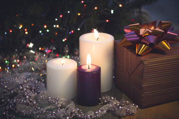 Obraz na płótnie Canvas A festive still life with a purple and two white candles of different size, a brown striped gift box with a dark shiny bow and a garland of beads and a ribbon. Dark Christmas background. Blurred bokeh