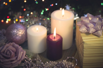 Obraz na płótnie Canvas A romantic festive still life with burning candles of different size, a beige striped gift box, a garland, a paper rose and a Christmas tree ball with glitter. Light purple and lilac colors. Dark