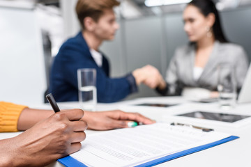 cropped shot of african american woman signing contract while blurred business partners shaking hands on background