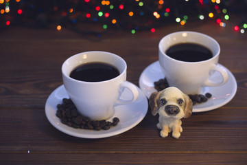 A festive still life with two white porcelian cups of hot black coffee. Coffee beans scattered on the saucers. A toy puppy in a close up. A rustic wooden background with Christmas lights. Dark
