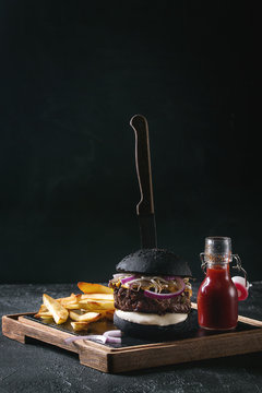 Homemade black bun hamburger with beef, mozzarella cheese, sprouts served on wooden slate serving board with french fries, knife and ketchup sauce on dark texture background.