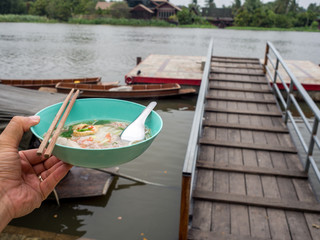 Left hand holding a bowl of noodles. The river is behind and there are noodle boats. thailand lifestyle