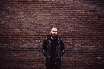 Fototapeta na wymiar Modern young bearded man in black style clothes posing against brick wall.