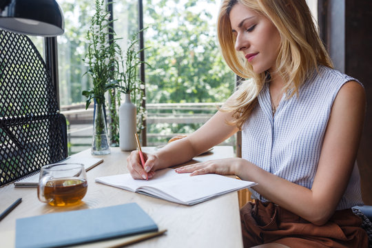 Pretty blonde Caucasian woman writing in her notebook at home office.