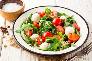 Fresh salad with arugula, cherry tomatoes, mozzarella cheese and walnuts on white wooden background.