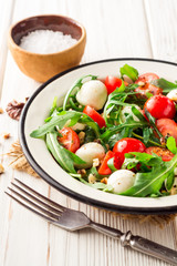 Fresh salad with arugula, cherry tomatoes, mozzarella cheese and walnuts on white wooden background.