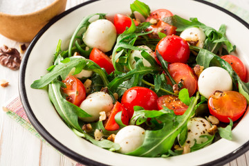 Fresh salad with arugula, cherry tomatoes, mozzarella cheese and walnuts on white wooden background.