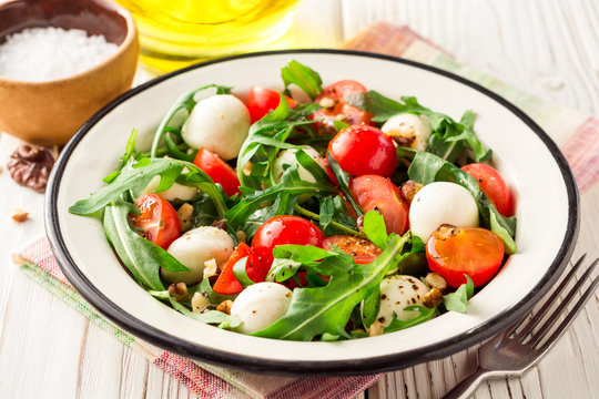 Fresh salad with arugula, cherry tomatoes, mozzarella cheese and walnuts on white wooden background.