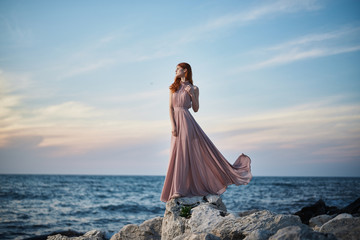 Beautiful young woman in a long dress standing by the sea, rocks