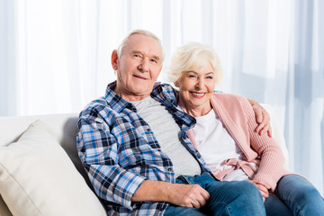 portrait of smiling senior wife and husband looking at camera while resting on sofa at home
