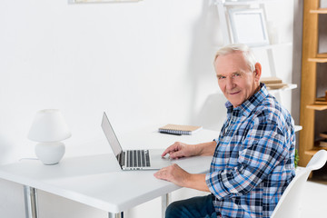 side view of senior man sitting at table with laptop and looking at camera at home