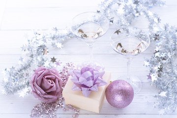 A romantic festive afternoon with two glasses of chapmagne, a beige gift box with a lilac shiny bow. Lilac rose, beads and a ball,a silver shiny garland on a white wooden table. Top view.