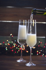 A festive still life with two wine glasses on a dark colored wooden background. Champagne is being poured into a glass from a bottle. Dark colorful bokeh