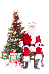 Portrait of Santa Claus sitting in chair next to Christmas tree, heap of gift boxes near him