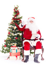 Portrait of Santa Claus sitting in chair next to Christmas tree, heap of gift boxes near him