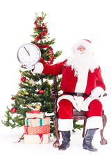 Portrait of Santa Claus sitting in chair next to Christmas tree holding clock, heap of giftboxes near him