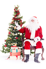 Portrait of Santa Claus sitting in chair next to Christmas tree, heap of gift boxes near him