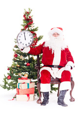 Portrait of Santa Claus sitting in chair next to Christmas tree holding clock, heap of giftboxes near him