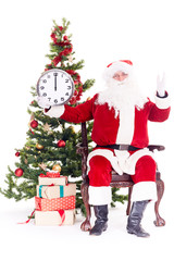 Portrait of Santa Claus sitting in chair next to Christmas tree holding clock, heap of giftboxes near him
