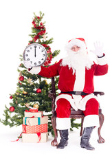 Portrait of Santa Claus sitting in chair next to Christmas tree holding clock, heap of giftboxes near him