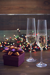 Two glasses of champagne and a burgundy gift box with a green and purple shiny bow on a table.Dark colored wooden background. Christmas fairy lights on the back. Dark colorful bokeh