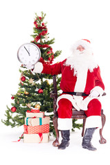 Portrait of Santa Claus sitting in chair next to Christmas tree holding clock, heap of giftboxes near him