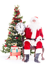 Portrait of Santa Claus sitting in chair next to Christmas tree holding clock, heap of giftboxes near him