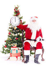 Portrait of Santa Claus sitting in chair next to Christmas tree holding clock, heap of giftboxes near him