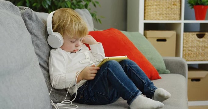 Cute little boy in the big white headphones listening to the music on his tablet device in the cozy kiving room. inside
